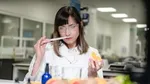 A woman in a lab coat and safety googles smells a paper-smelling strip while holding an orange  in her other hand.