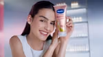 A white woman with dark hair and glowing skin, holding a tube of Vaseline Gluta Hya Dewy Radiance cream. Pink, premium packaging.