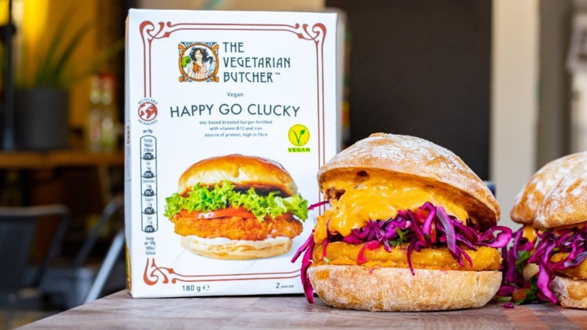 Two well garnished vegan chicken burgers, next to a packet of The Vegetarian Butcher Happy Go Clucky vegan breaded burgers.