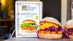 Two well garnished vegan chicken burgers, next to a packet of The Vegetarian Butcher Happy Go Clucky vegan breaded burgers.