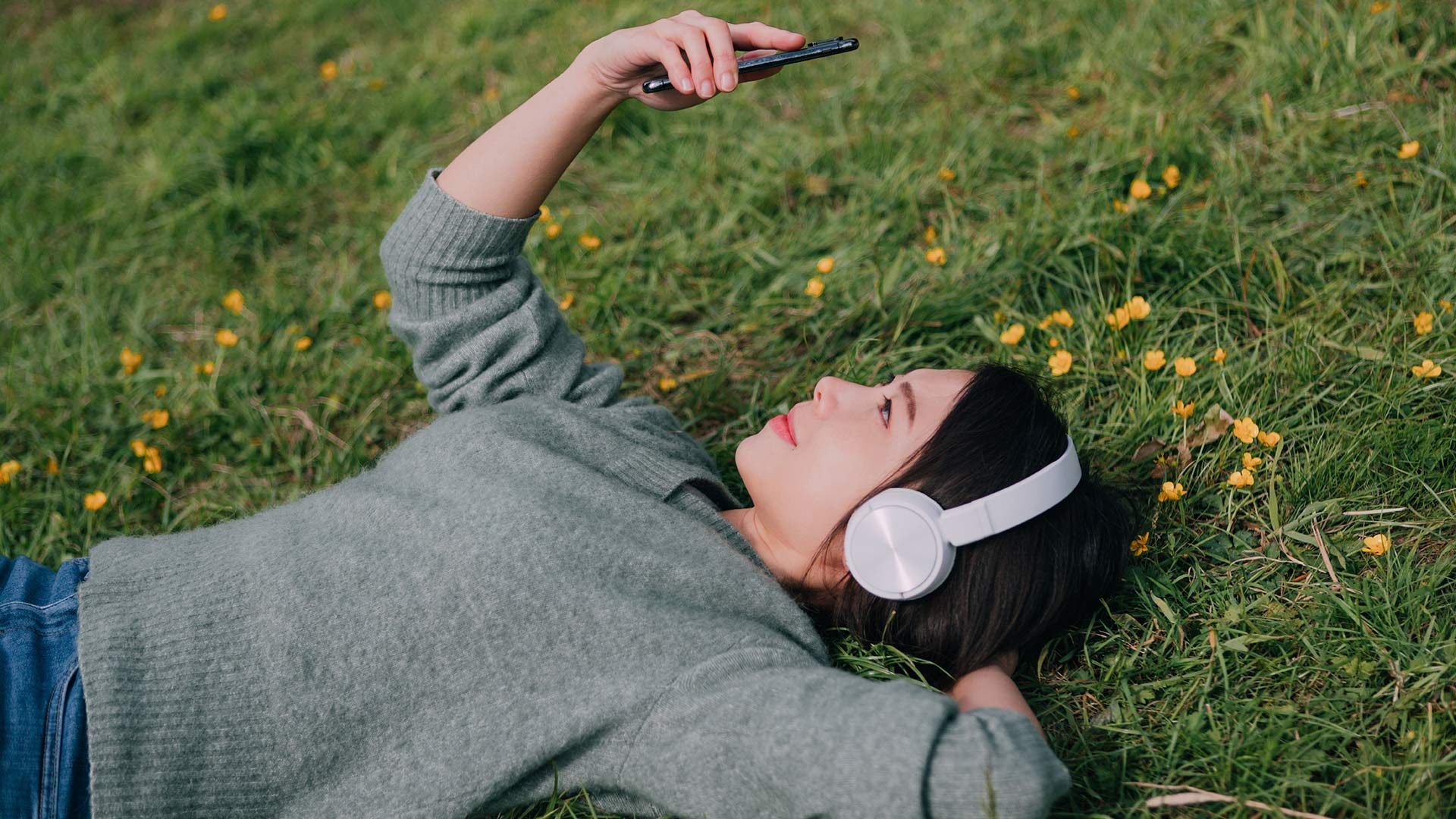 Young woman wearing headphones and studying phone, relaxes on a grassy field