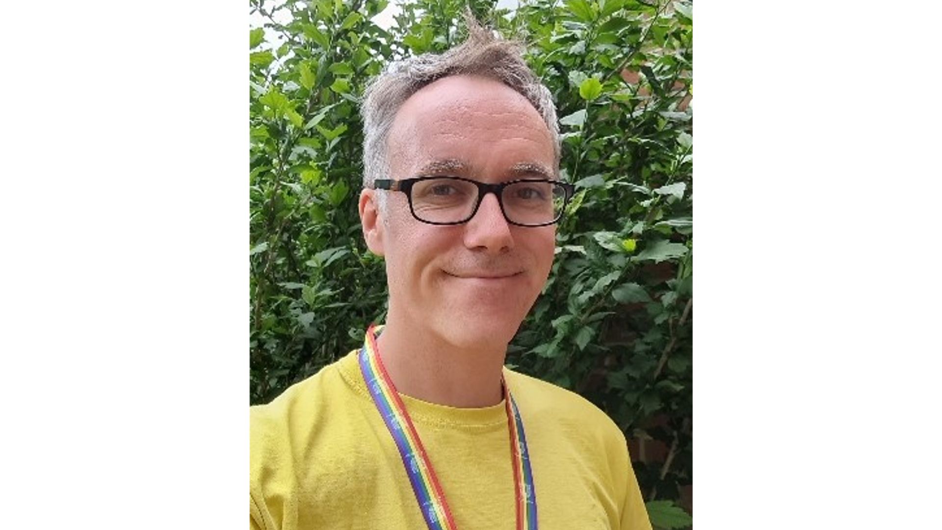 David smiling at camera wearing glasses, a yellow t-shirt and a rainbow lanyard