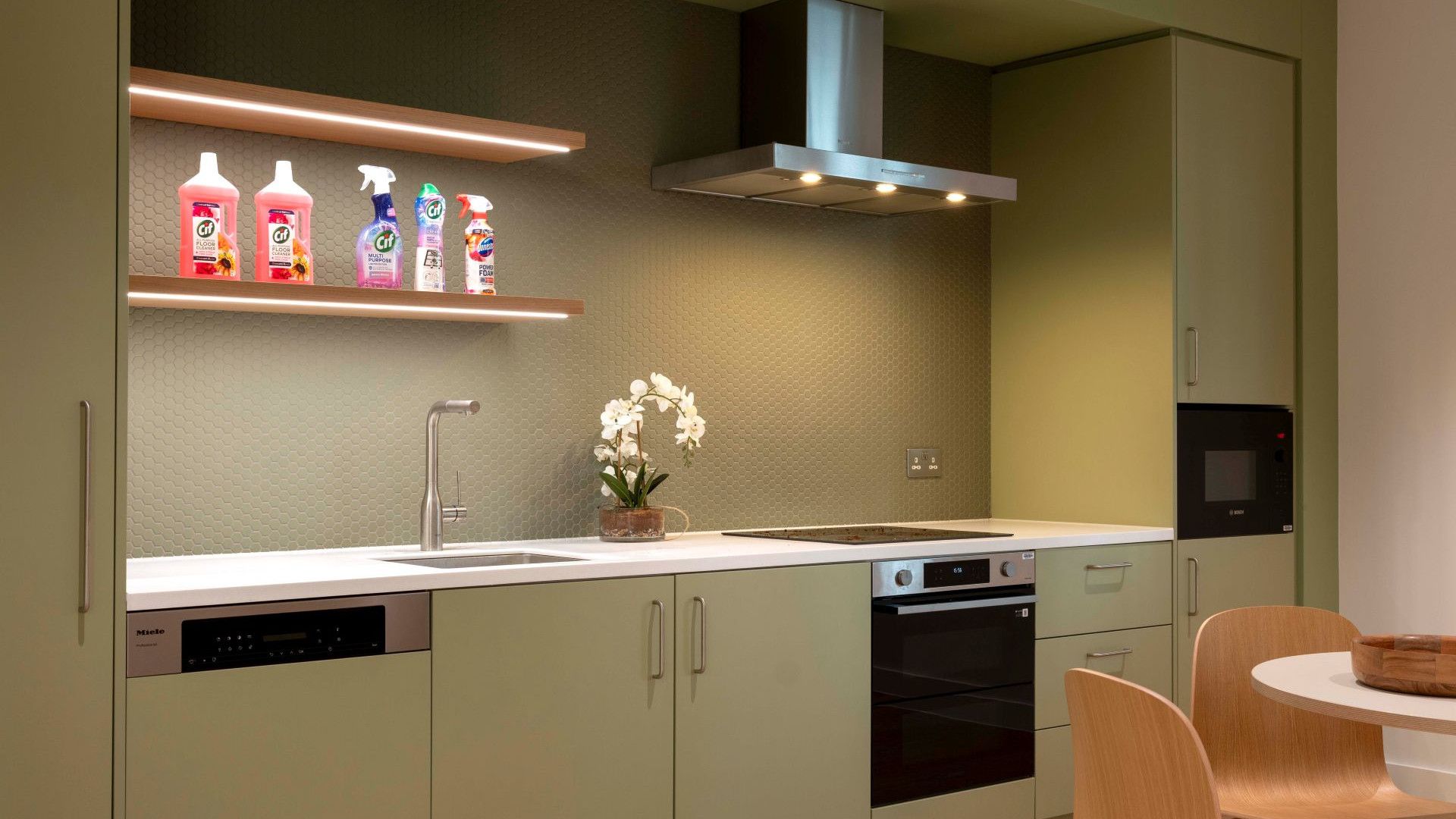 Modern fitted kitchen with green cabinets, integrated appliances and cleaning products displayed on a shelf.