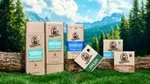 Dr. Squatch grooming products – including shampoo, conditioner, deodorant, and soaps – arranged on logs and grass in a natural outdoor setting with trees, mountains, and a blue sky in the background.