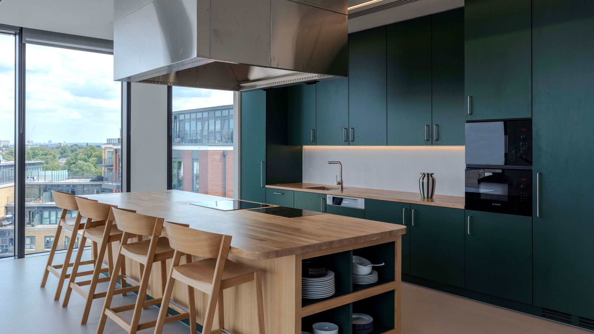 Modern office kitchen with dark green cabinets, a wooden island, bar stools and floor-to-ceiling windows.