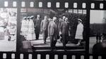 Historic filmstrip shows suited officials touring a facility as uniformed staff stand in line.